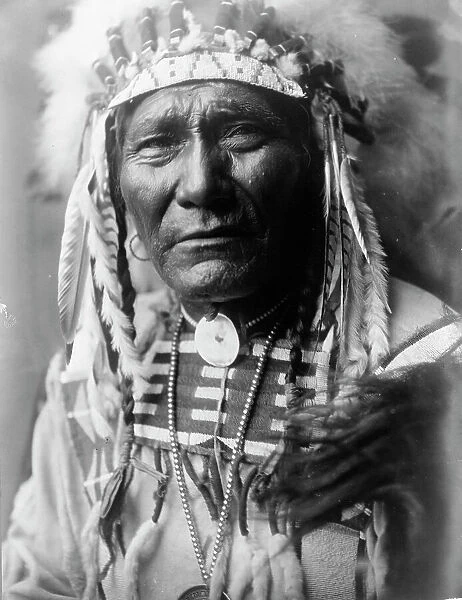 Ghost Bear, Crow Indian, Montana, head-and-shoulders portrait, facing front, feather... c1908. Creator: Edward Sheriff Curtis