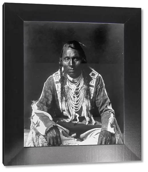 Wades in Water, Piegan Indian, full-length portrait, seated on floor, facing front... c1910. Creator: Edward Sheriff Curtis