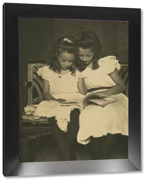 Two girls wearing white dresses and dark stockings reading a book, c1900. Creator: Virginia M Prall