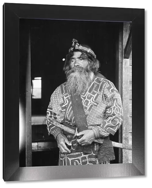 Ainu chief wearing a headdress and holding a sword standing in a doorway, 1908. Creator: Arnold Genthe