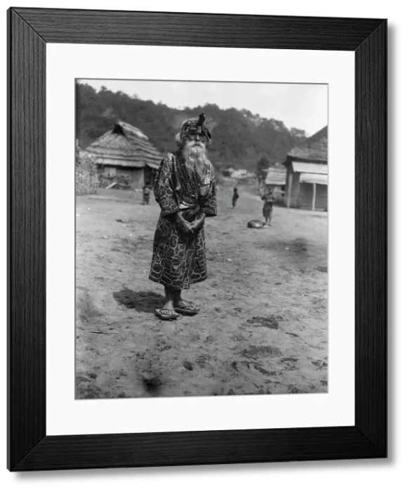 Framed Print : Ainu chief wearing a headdress standing in the village lane, 1908. Creator: Arnold Genthe