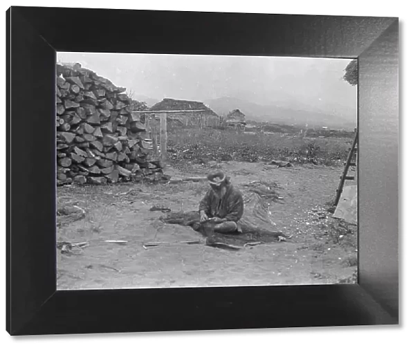 Ainu man seated outside working on nets, 1908. Creator: Arnold Genthe