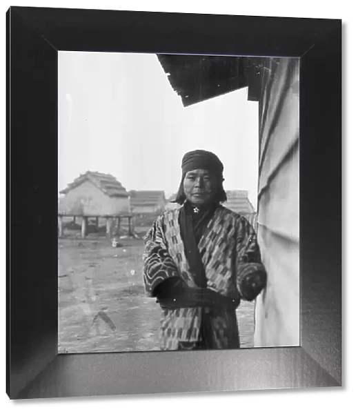 Ainu standing outside by the wall of a wooden hut, 1908. Creator: Arnold Genthe