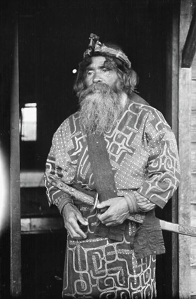 Ainu chief wearing a headdress and holding a sword standing in a doorway, 1908. Creator: Arnold Genthe