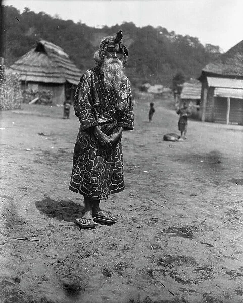 Ainu chief wearing a headdress standing in the village lane, 1908. Creator: Arnold Genthe