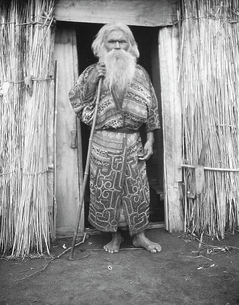 Ainu man holding a staff standing at the doorway of a hut, 1908. Creator: Arnold Genthe