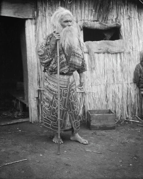 Ainu man holding a staff standing outside a hut, 1908. Creator: Arnold Genthe
