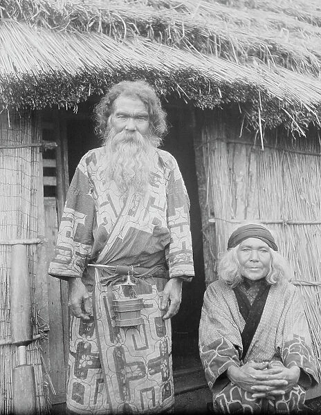 Ainu man and seated woman at the entrance of a hut, 1908. Creator: Arnold Genthe