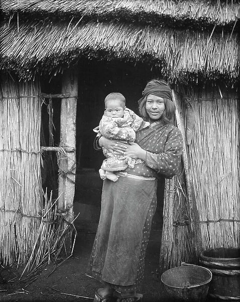Ainu woman holding a child standing outside a hut, 1908. Creator: Arnold Genthe