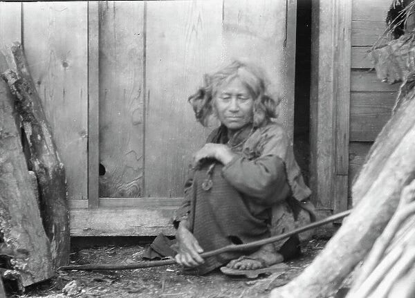 Ainu woman seated by the door of a wooden hut, 1908. Creator: Arnold Genthe