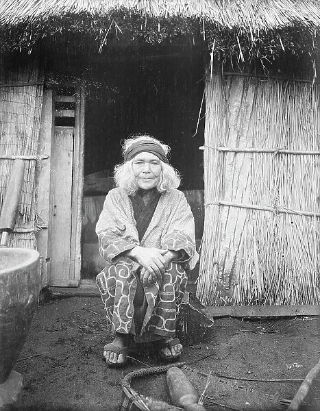 Ainu woman seated in front of the doorway of a hut, 1908. Creator: Arnold Genthe