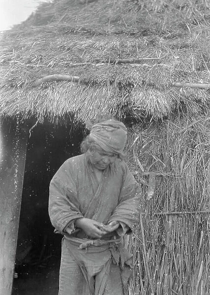 Ainu woman standing outside the entrance of a hut, 1908. Creator: Arnold Genthe