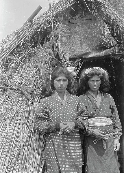 Two Ainu women standing at the entrance of a hut, 1908. Creator: Arnold Genthe
