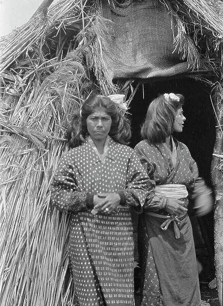 Two Ainu women standing outside the entrance of a hut, 1908. Creator: Arnold Genthe