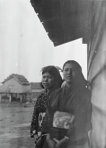 Two Ainu women standing outside by the wall of a wooden hut, 1908. Creator: Arnold Genthe