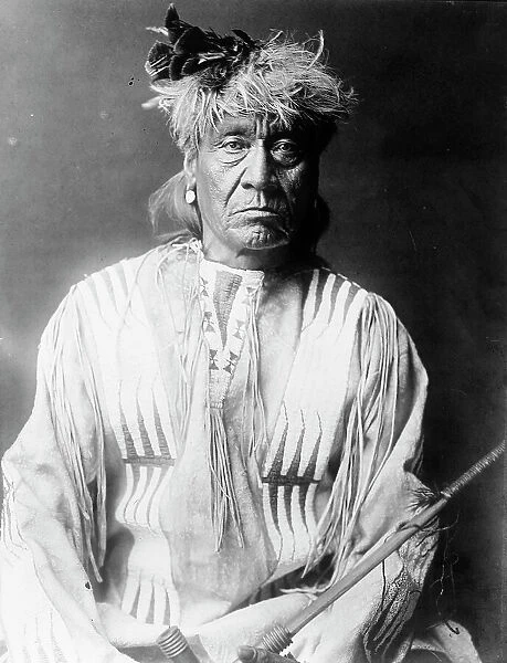 Atsina Indian, Red Whip, half-length portrait, seated, facing front, wearing feather... c1908. Creator: Edward Sheriff Curtis