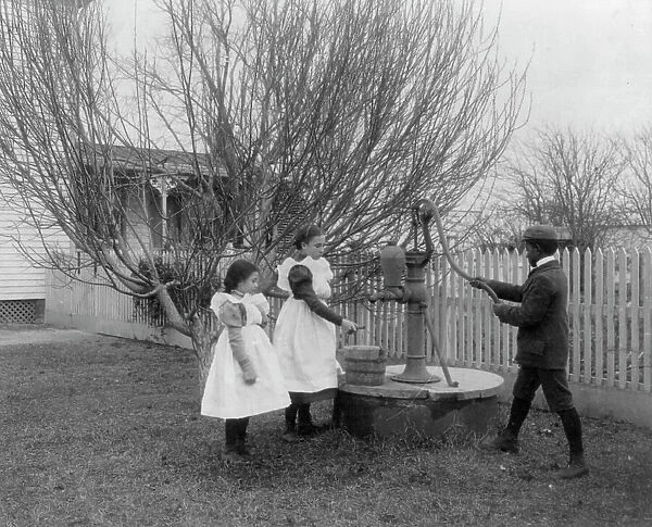 Two girls and a boy pumping water at well of Hampton Institute graduate, 1899 or 1900. Creator: Frances Benjamin Johnston