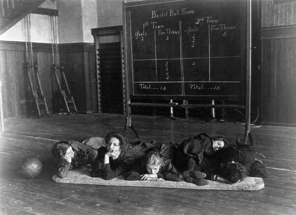 Five girls lying on mat in gymnasium in front of basketball scoreboard... Washington, D.C. (1899?) Creator: Frances Benjamin Johnston