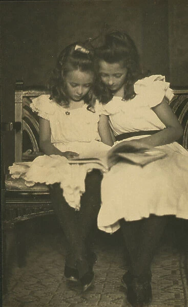 Two girls wearing white dresses and dark stockings reading a book, c1900. Creator: Virginia M Prall