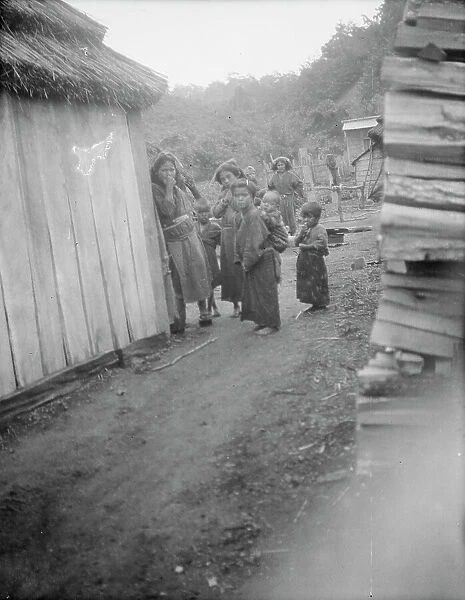 Group of Ainu children standing in a passageway between huts, 1908. Creator: Arnold Genthe