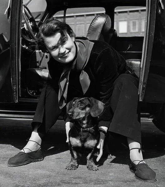 Photographer Kerstin Bernhard sits in the doorway of her car and holds a dachshund, 1955-1960. Creator: Unknown