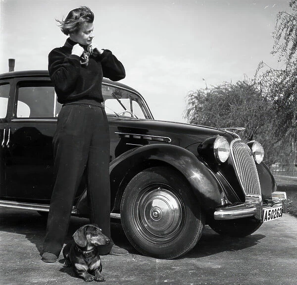 Photographer Kerstin Bernhard stands next to a car with her dachshund, 1955-1960. Creator: Unknown