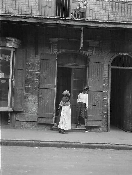 View from across street of a man and woman standing by a doorway and a woman seated...c1920-1926. Creator: Arnold Genthe