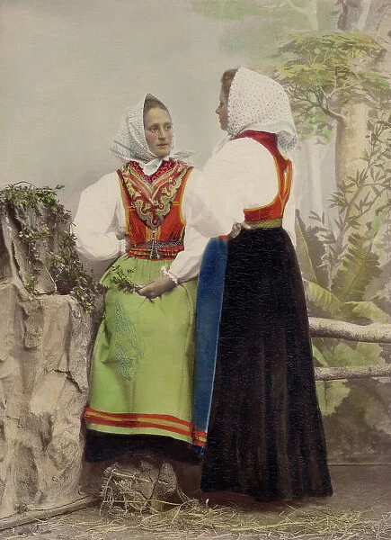 Two young women pose in folk costumes, with dotted headdresses, 1886-1890. Creator: Helene Edlund