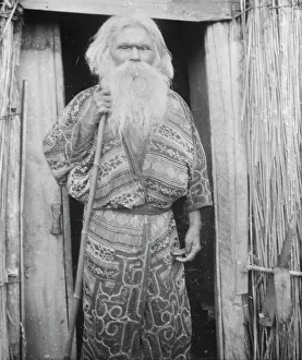 Ainu man holding a staff standing at the doorway of a hut, 1908. Creator: Arnold Genthe