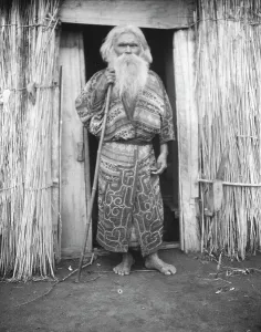 Aino Prints: Ainu man holding a staff standing at the doorway of a hut, 1908. Creator: Arnold Genthe