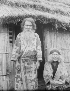 Aino Prints: Ainu man and seated woman at the entrance of a hut, 1908. Creator: Arnold Genthe