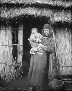 Aino Prints: Ainu woman holding a child standing outside a hut, 1908. Creator: Arnold Genthe
