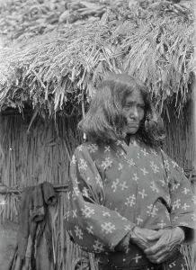 Aino Prints: Ainu woman standing outside a hut, 1908. Creator: Arnold Genthe