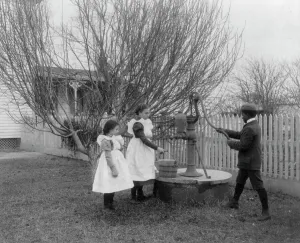Pumping Prints: Two girls and a boy pumping water at well of Hampton Institute graduate