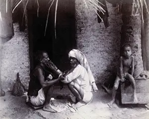 Sitting On Ground Prints: Two Indian men crouching; one is cutting the other's hair, while a girl sits.. c1890s