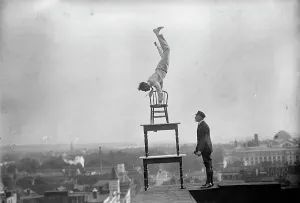 Publicity Stunt Prints: J. Reynolds, Performing Acrobatic And Balancing Acts On High Cornice Above 9th Street