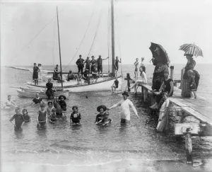 Men, women, and children in bathing suits on dock, sailboats, and in water... c1880. Creator: Unknown