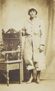 Photographic postcard of a woman standing next to a wooden chair, 1918-1930 Photographic postcard of a woman standing next to a wooden chair, 1918-1930