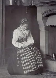 Edlund Helene Prints: A woman sits in front of a fireplace and reads dressed in a Dalarna costume